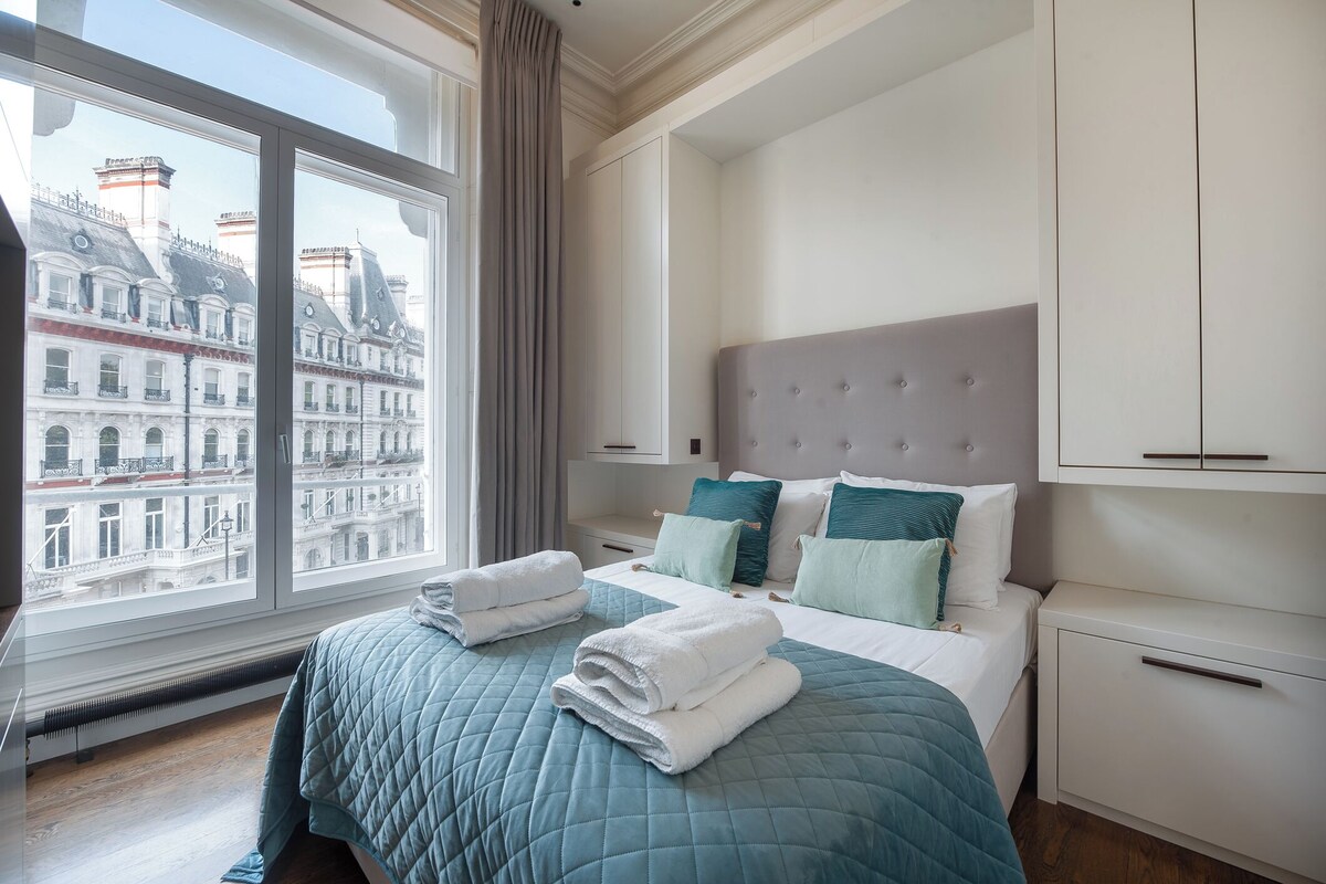 Ein Hotelzimmer mit großem Fenster, einem Bett mit gesteppter blauer Bettdecke und Blick auf ein Gebäude gegenüber.