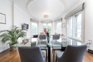 A spacious room with a glass table, grey chairs, a potted plant, and large windows offering a view of buildings.