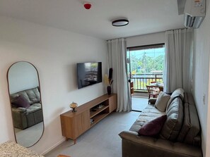 A modern living room with a flat-screen TV, a sofa, a coffee table, and a balcony with a view.