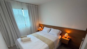A hotel room with a neatly made bed, two bedside lamps, a wooden headboard, and a window with curtains.