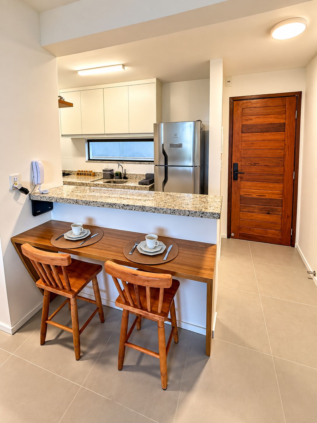 A compact kitchen with a built-in refrigerator, a microwave, and a coffee machine. There are two wooden chairs and a small table with a cup and saucer.