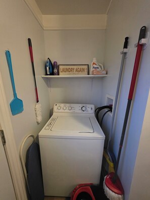 A laundry room with a washing machine, a mop, a broom, and a shelf with laundry supplies.