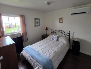Quarto com cama, mesa de cabeceira, cômoda, janela com cortinas e um aparelho de ar condicionado.