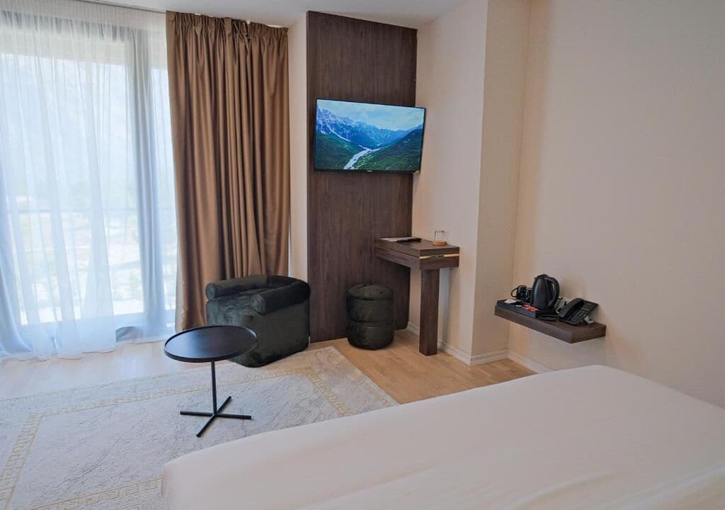A hotel room with a bed, a small round table, a sofa, a TV mounted on the wall, a coffee machine, and a shelf with a phone and a remote control.