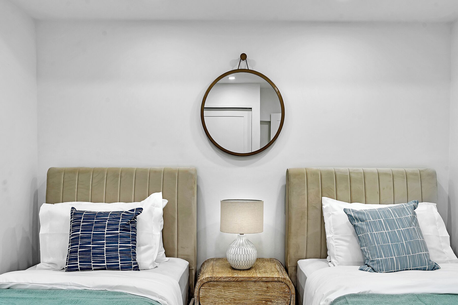 Quarto de hotel com duas camas, um espelho redondo, um abajur e uma pequena mesa de cabeceira de madeira.