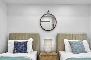 Hotellihuone, jossa on kaksi sänkyä, pyöreä peili, yöpöytälamppu ja pieni puinen sivupöytä.