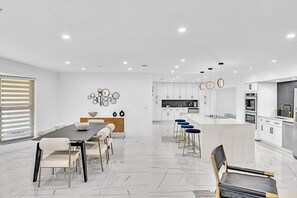 A modern kitchen with white cabinetry, a central island with bar stools, and a dining area with a long table and chairs.