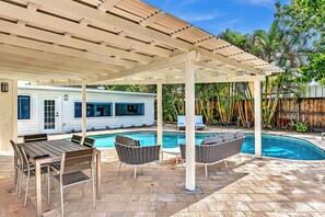 A covered outdoor area with a pool, patio furniture, and a house in the background.
