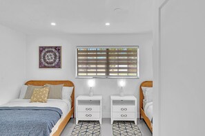 A bedroom with two beds, a window with blinds, a wall-mounted artwork, and bedside tables with drawers.