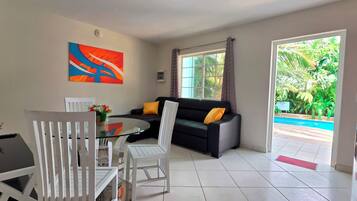 A modern living room with a glass table, white chairs, a black sofa with yellow cushions, and a colorful abstract painting on the wall.