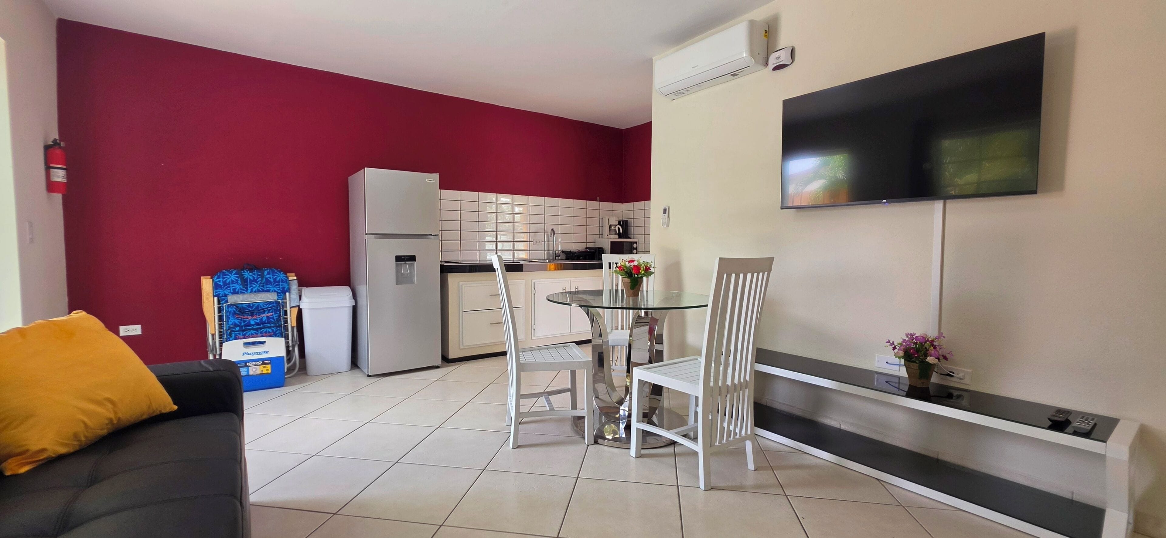 A modern living room with a sofa, a dining table with chairs, a kitchen area with a refrigerator, and a mounted television.