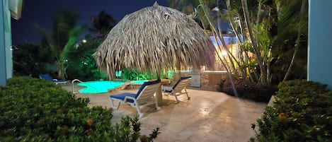 A pool area with lounge chairs and a thatched umbrella, surrounded by palm trees and greenery.