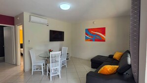 A living room with a black sofa, white chairs, a TV, and a painting. - Cozy 1-bedroom Apartment 3 km From Eagle Beach (Noord)