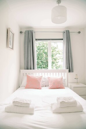 Ein Schlafzimmer mit Bett, Kissen, Nachttisch, Lampe und einem Fenster mit Vorhängen.