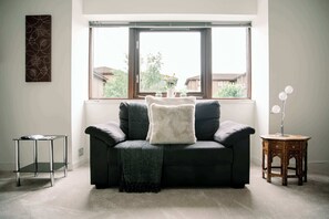 Ein modernes Wohnzimmer mit einem dunklen Sofa, einem kleinen Beistelltisch und einem Fenster mit Blick auf Häuser.