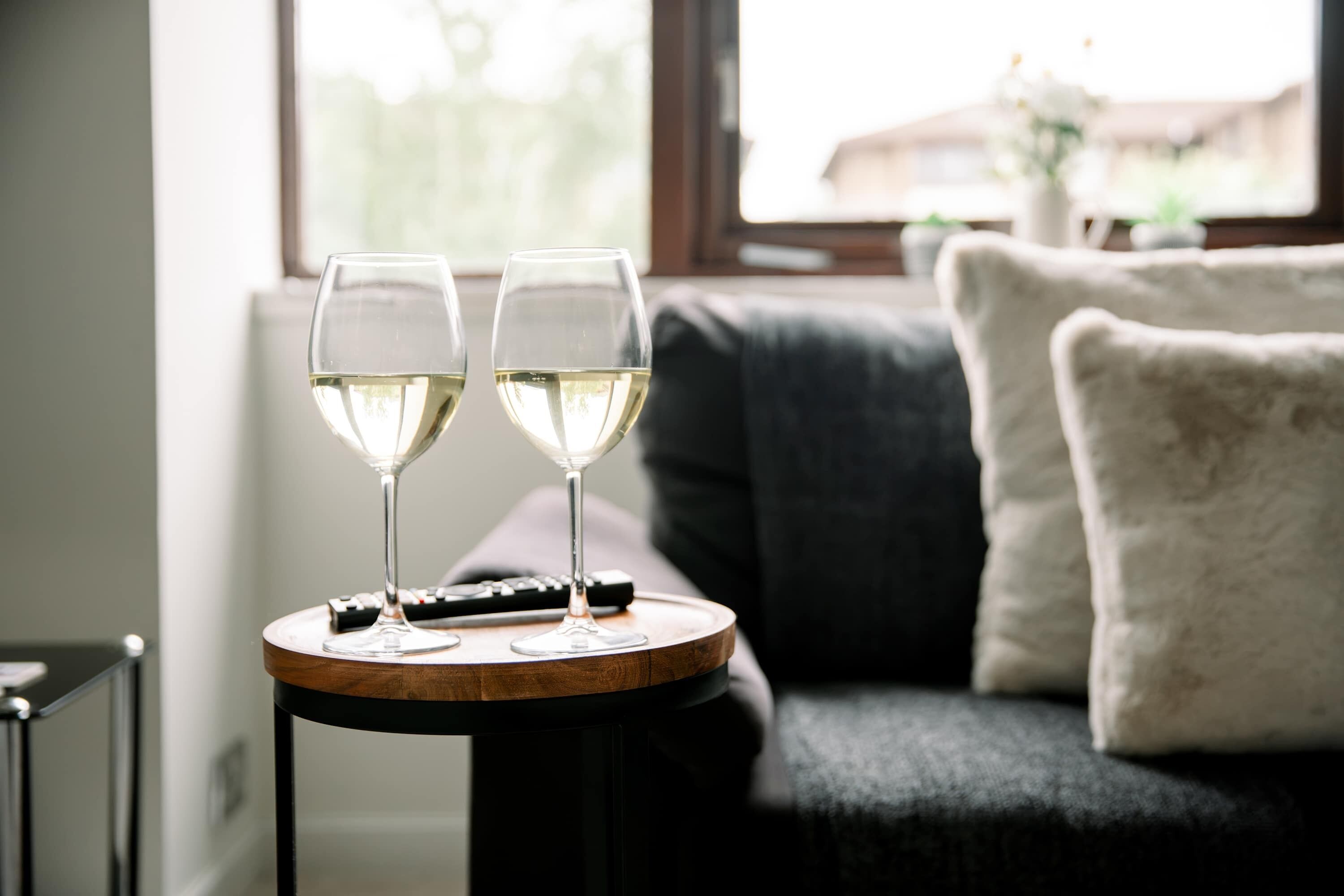 Two wine glasses on a round wooden table with a remote control beside them, placed on a dark sofa with a white pillow in a room with a window.