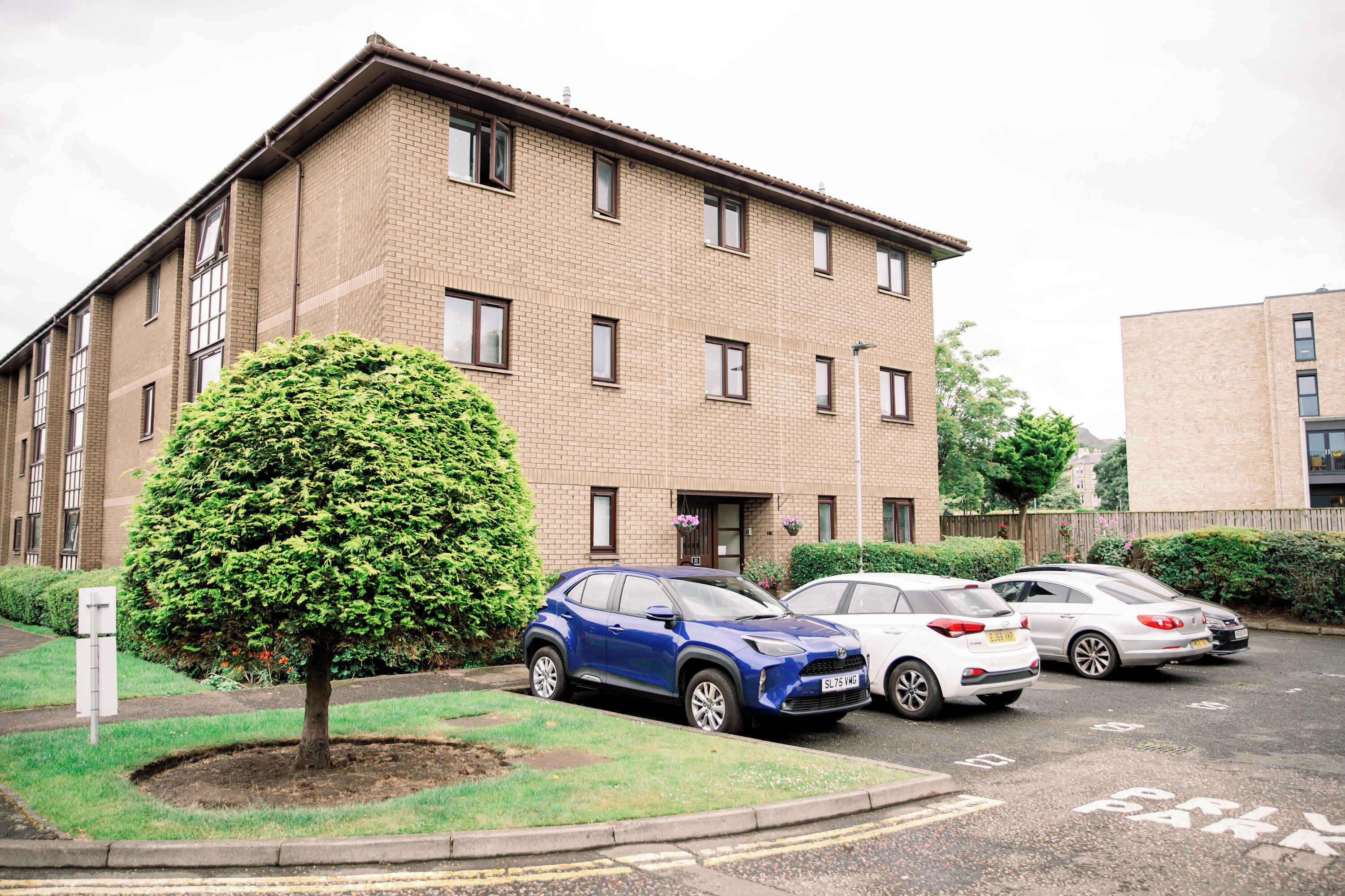 Un edificio residenziale a più piani con facciata in mattoni, un piccolo albero nel giardino antistante e diverse auto parcheggiate.