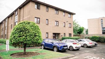 Un edificio residenziale a più piani con facciata in mattoni, un piccolo albero nel giardino antistante e diverse auto parcheggiate.