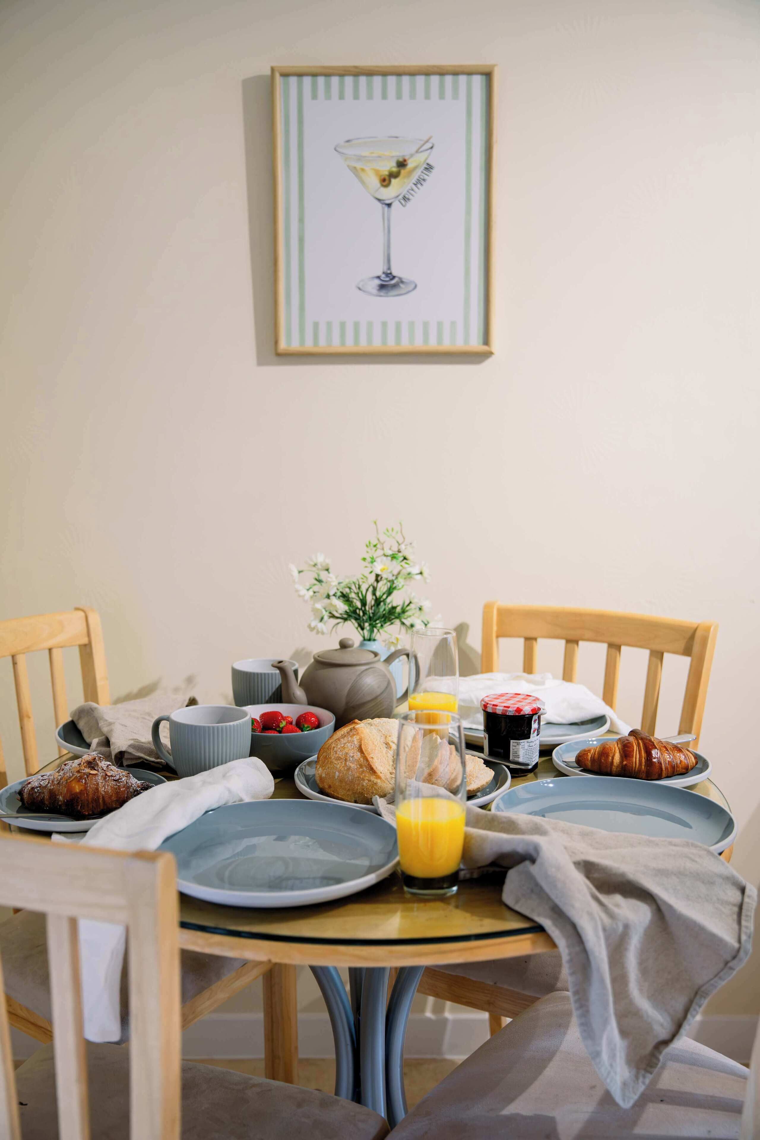A breakfast setting with a table, chairs, a teapot, cups, a plate with strawberries, a glass of orange juice, and a croissant.