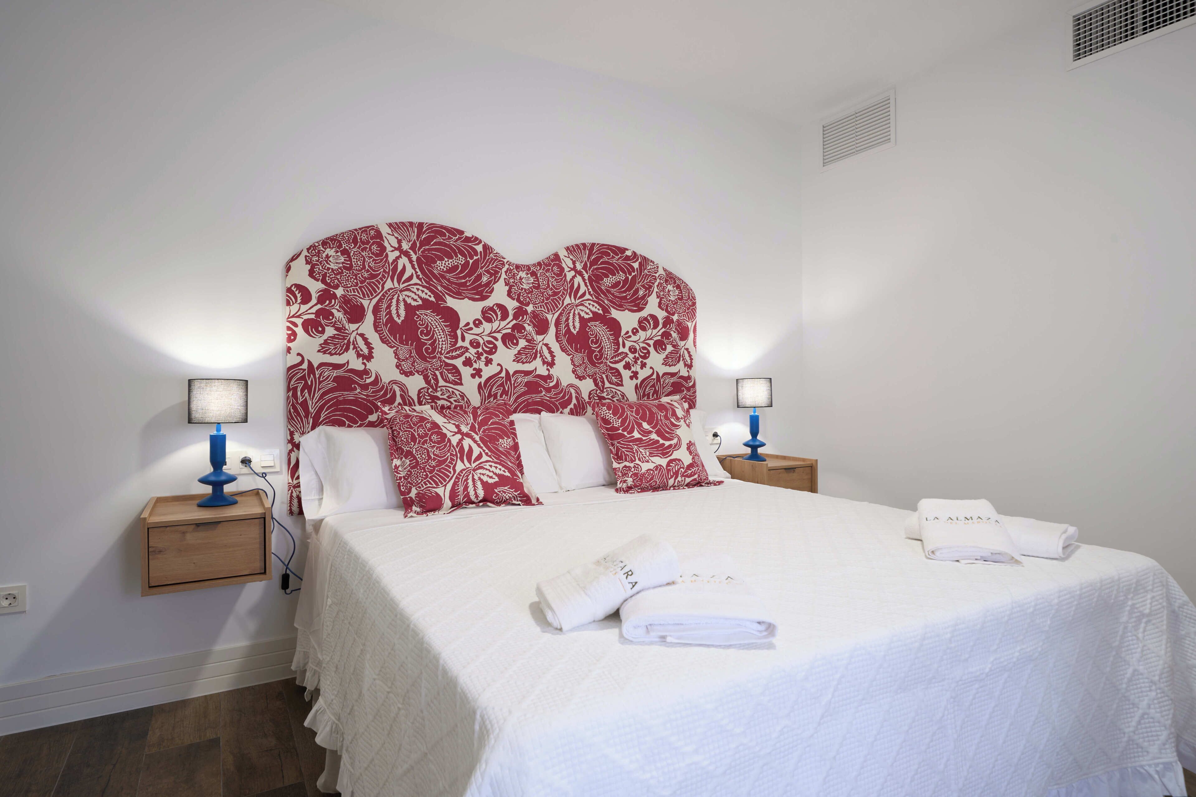 Een hotelkamer met een groot bed, twee nachtkastjes met lampen, een hoofdeinde met bloemenprint en een witte handdoek op het bed.