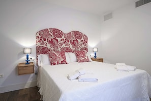 Habitación de hotel con una cama grande, dos mesitas de noche con lámparas, un cabecero con estampado floral y una toalla blanca sobre la cama.