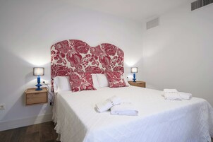 Habitación de hotel con una cama grande, dos mesitas de noche con lámparas, un cabecero con estampado floral y una toalla blanca sobre la cama.