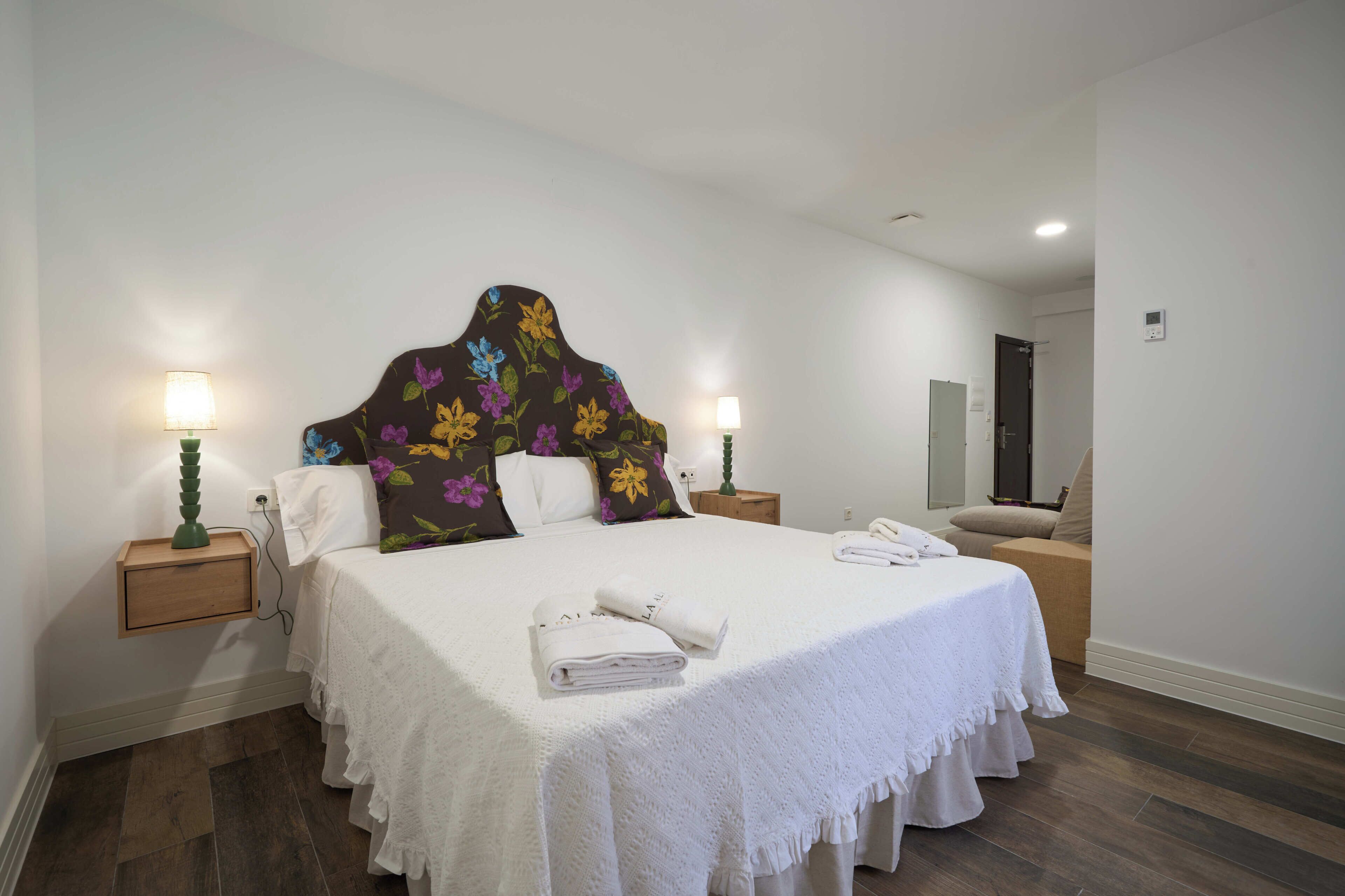 Una camera d'albergo con un letto grande, due comodini con lampade, un bagno visibile attraverso una porta aperta e un pavimento in legno.