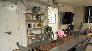 Un comedor con una mesa larga, sillas y una alfombra. Hay una estantería con diversos objetos y un televisor montado en la pared.