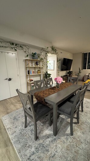 A dining area with a long table, chairs, and a rug. There is a bookshelf with various items and a television mounted on the wall. - Koreatown, LA -360 City DTLA Views- 2BD/2BTH w/Parking, Gym, Game Room (Commerce)