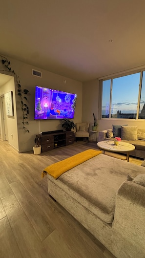 A modern living room with a flat-screen TV, a sofa, a coffee table, and a view through a large window. - Koreatown, LA -360 City DTLA Views- 2BD/2BTH w/Parking, Gym, Game Room (Commerce)
