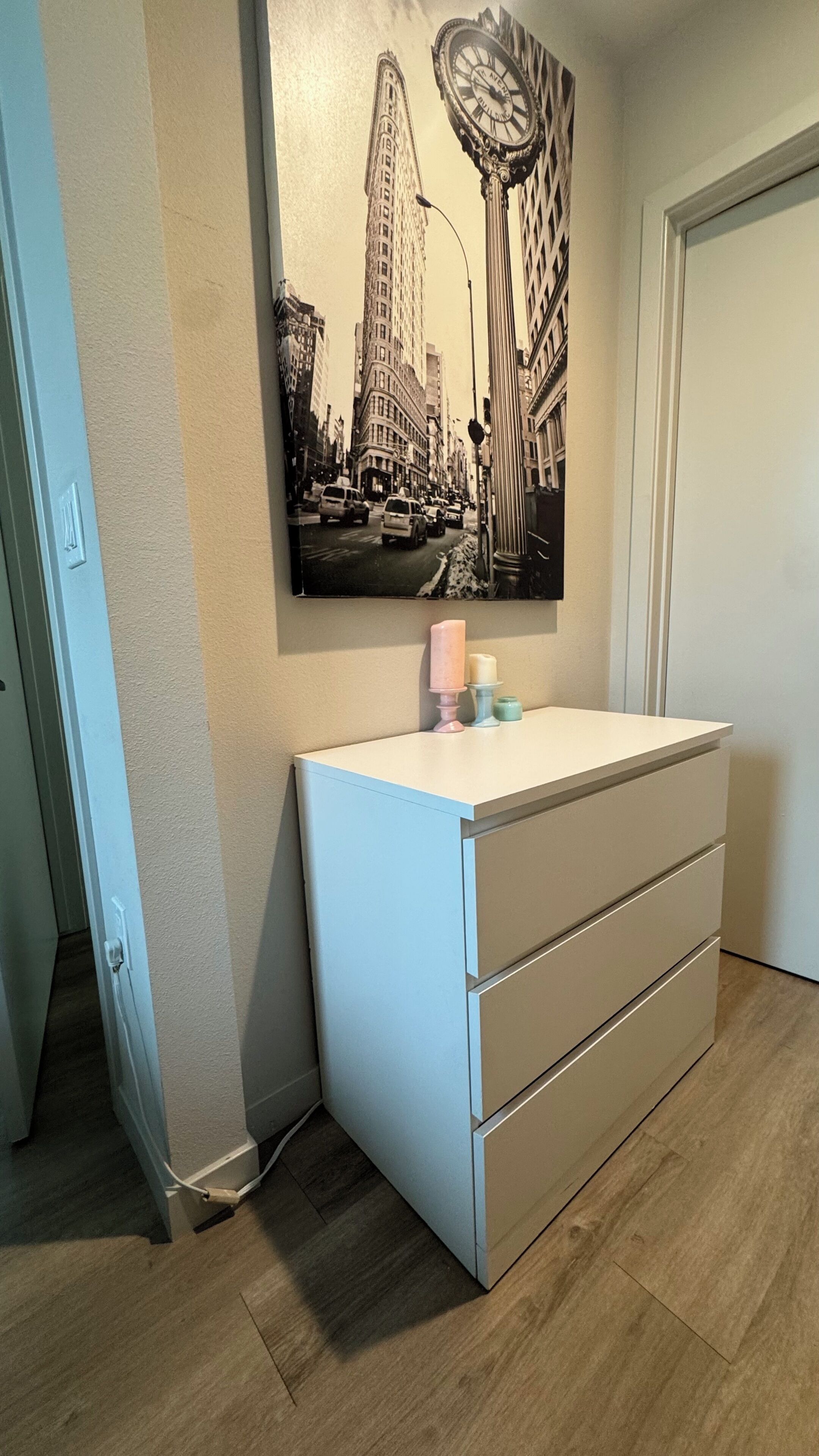 Huone, jossa on valkoinen lipasto, kaupunkimaisemaa esittävä maalaus ja ovi.