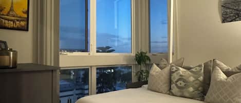 A bedroom with a large bed, a desk, and a window with a city view.