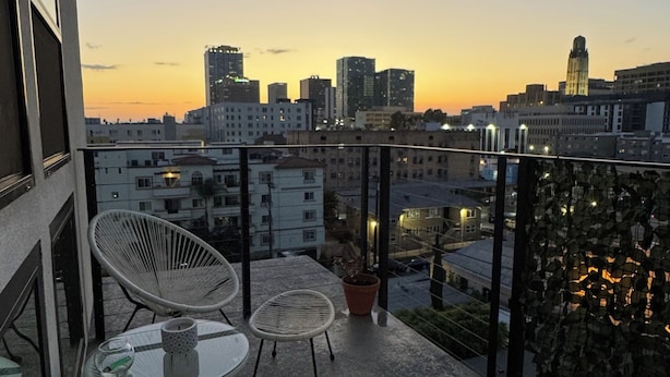 Un balcón con sillas y mesas de mimbre, con vistas a un paisaje urbano al atardecer.