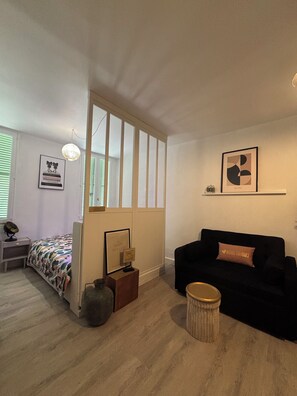 Ein Zimmer mit einem Bett, einem schwarzen Schlafsofa, einem kleinen runden Tisch, einer Wandregal und einem Fenster mit Jalousien.