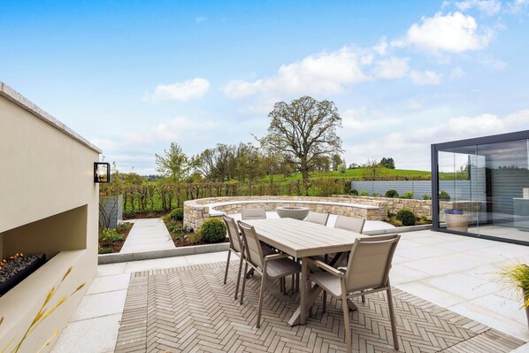An outdoor patio with a stone fire pit, a dining table with chairs, and a view of a lake and trees.