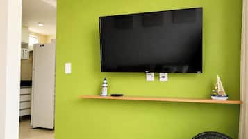 A room with a flat-screen TV mounted on a green wall, a small shelf with a decorative item, and a fan on the floor.