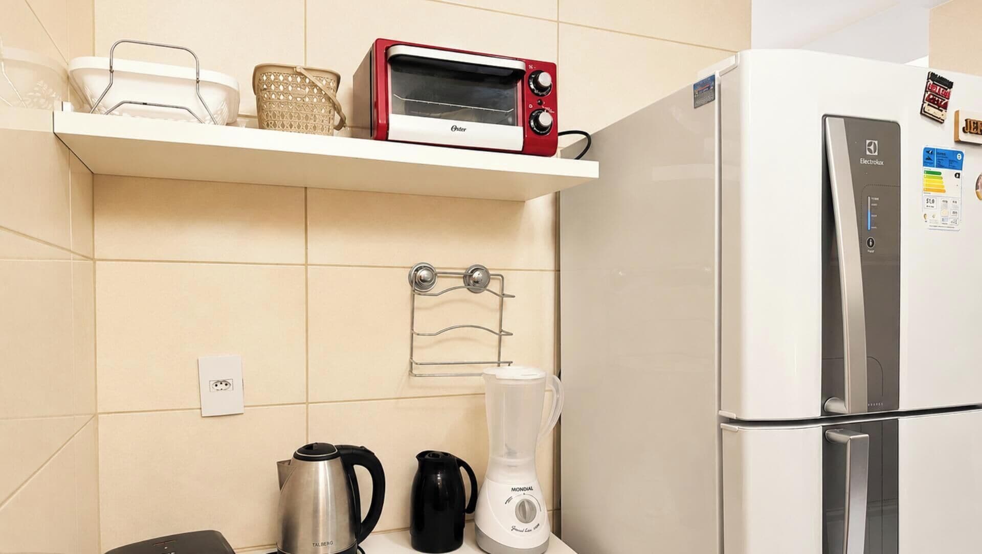 A kitchen with a red Oster toaster oven, a white refrigerator, a black kettle, a white blender, and a metal drying rack.
