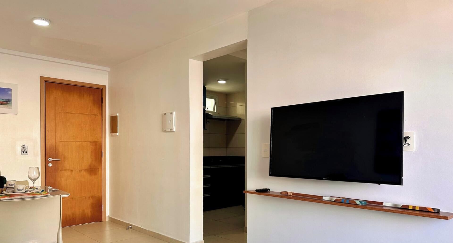 A modern interior with a wooden door, a flat-screen TV mounted on the wall, and a kitchen area visible through an open doorway.