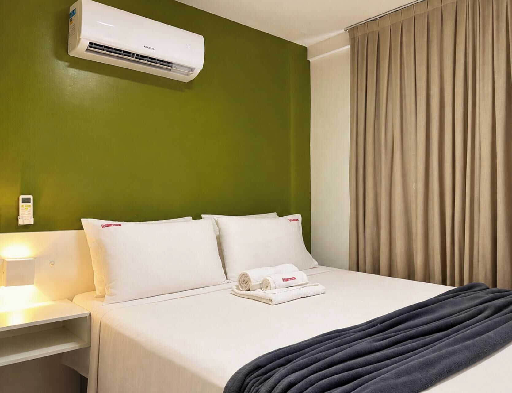 A hotel room with a bed, pillows, a blanket, an air conditioning unit, a bedside table, a lamp, and a curtain.