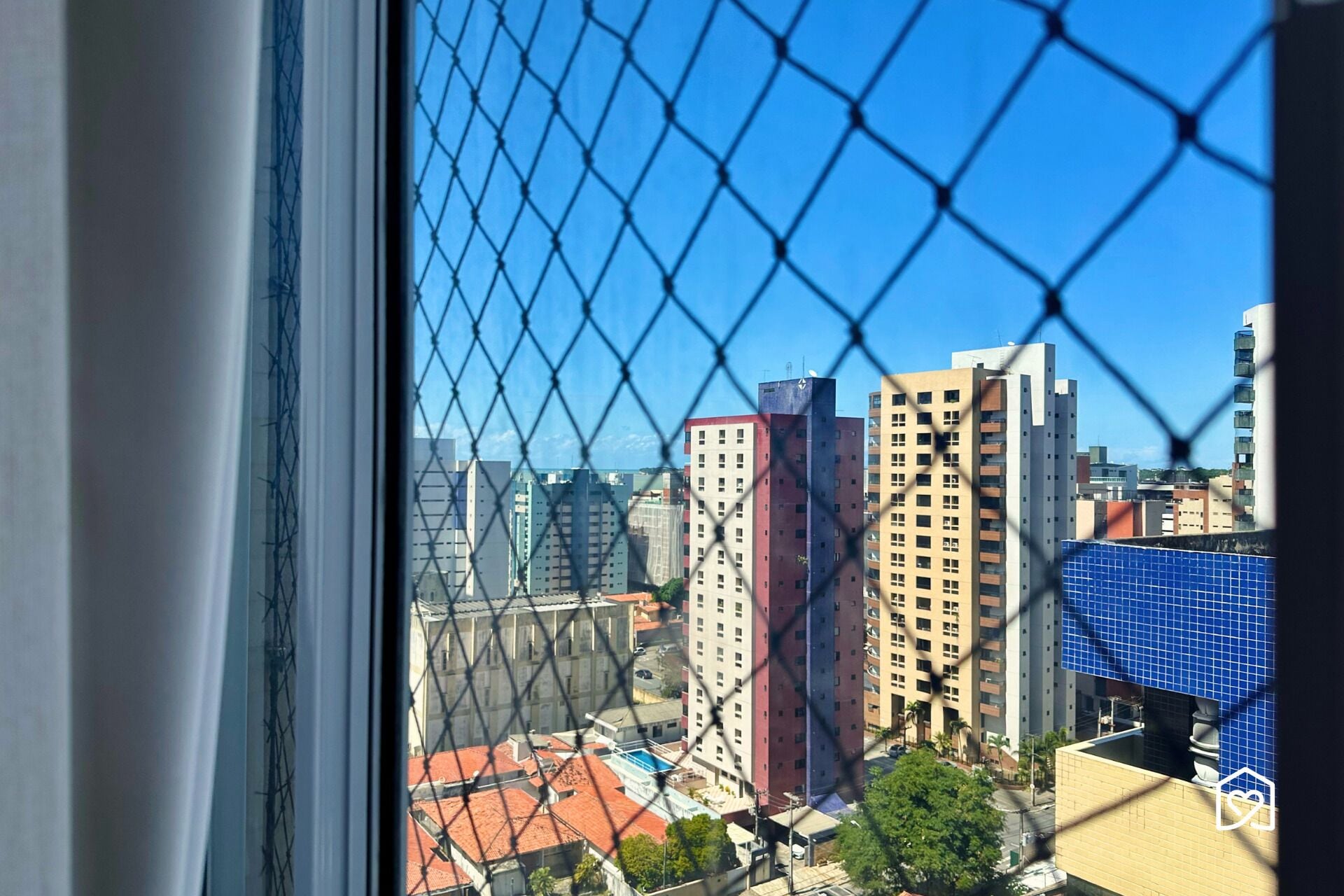 A view through a chain-link fence of a cityscape with high-rise buildings, including a red and white building and a blue building with a solar panel.