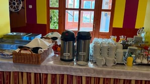 Montaje de bufé con dispensadores de agua y café, una cesta de frutas, y platos y vasos apilados.