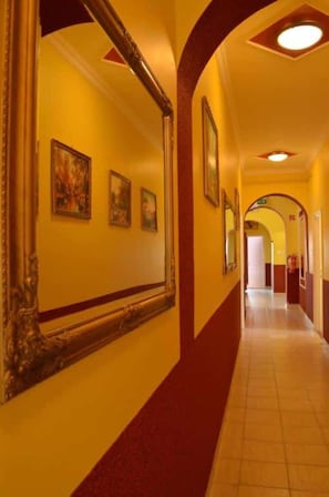 A corridor with yellow walls, red trim, and tiled flooring. There are framed pictures on the walls and a large mirror on the left. - Hotel Napsugár (Balatonmáriafürdő)