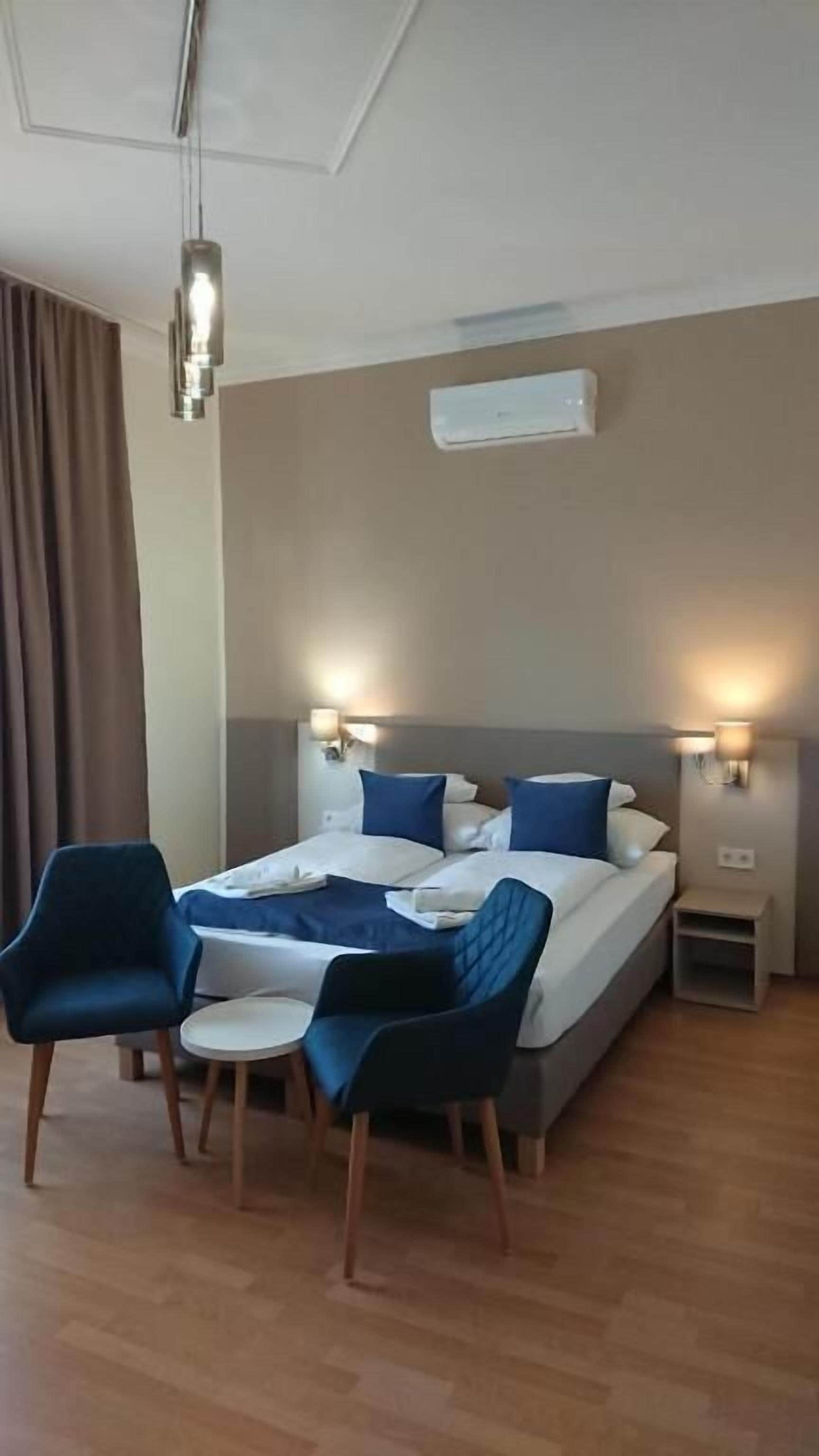 A hotel room with a bed, bedside tables, a chair, a small table, and a hanging light fixture.