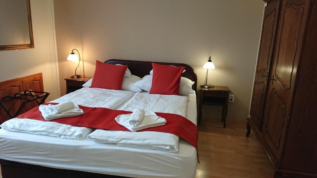 Una cama bien hecha con ropa de cama blanca y detalles rojos, dos lámparas de noche y un armario de madera.