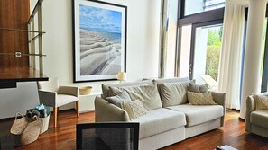A modern living room with a large window, a grey sofa, a small side table, and a framed picture of a desert landscape. - Troia Sea and Pool Townhouse (Grandola)