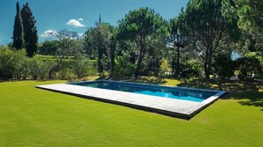 A rectangular swimming pool with a wooden deck, surrounded by a well-maintained lawn and various trees. - Troia Sea and Pool Townhouse (Grandola)