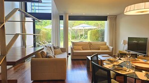 A modern living room with a sofa, a dining table set for two, a TV, and a staircase leading to an upper level. - Troia Sea and Pool Townhouse (Grandola)