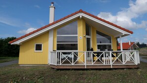 Uma casa amarela com telhado vermelho, chaminé e varanda com grades brancas.