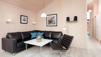 A modern living room with a black leather sofa, a white coffee table, and a black chair. There are framed pictures on the wall, a pendant light, and a shelf with speakers.