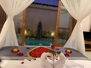 A bed with a canopy, a decorative flower arrangement, and a view of a swimming pool and statue. - One Bedroom Romantic  private with pool (ubud)